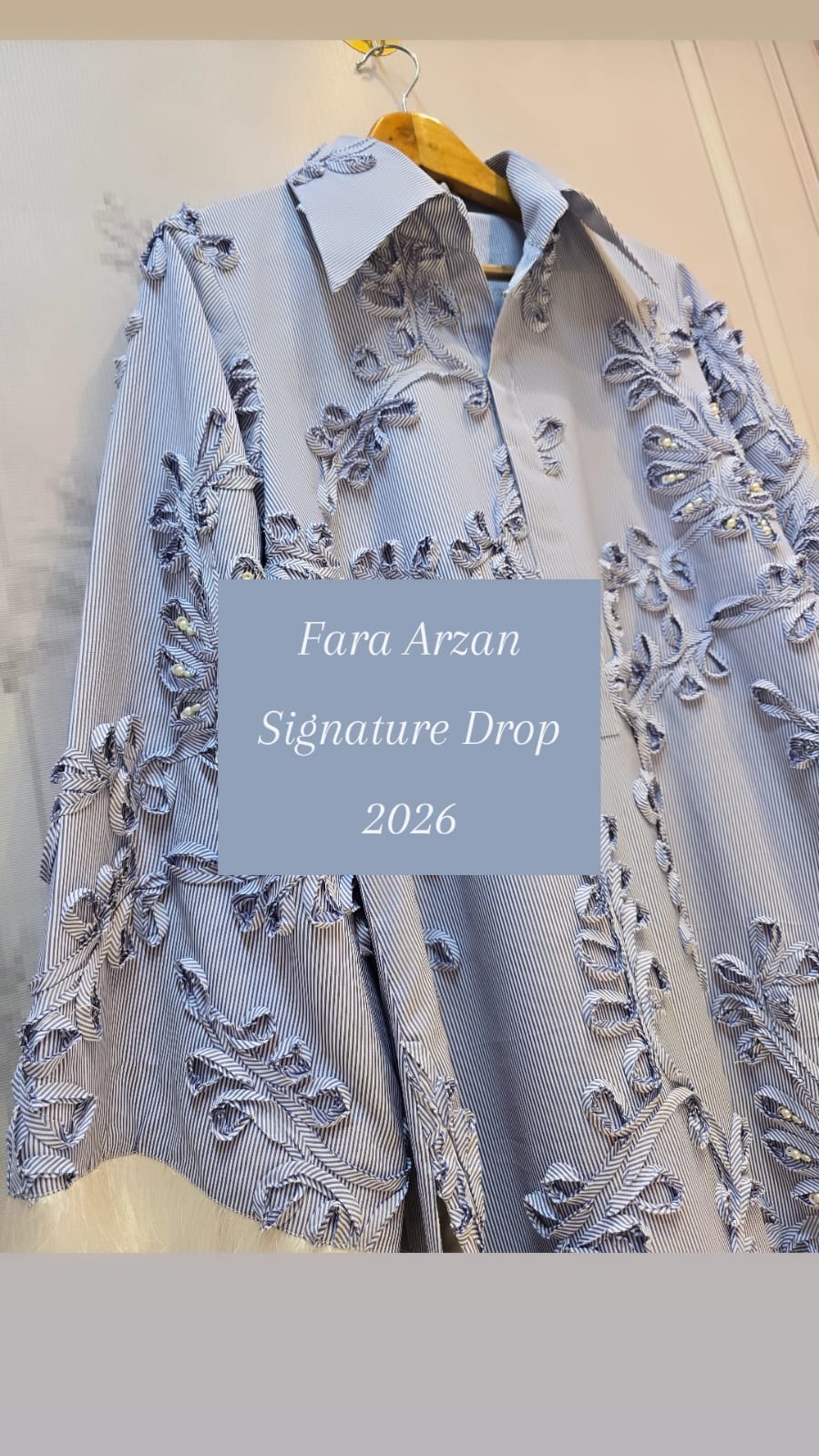 A light blue and white striped button-down shirt featuring heavy floral embroidery is displayed on a hanger.